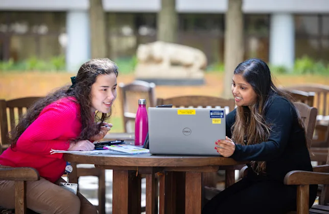 Students at an outdoor table look at a laptop with the Nittany Lion shrine in the background