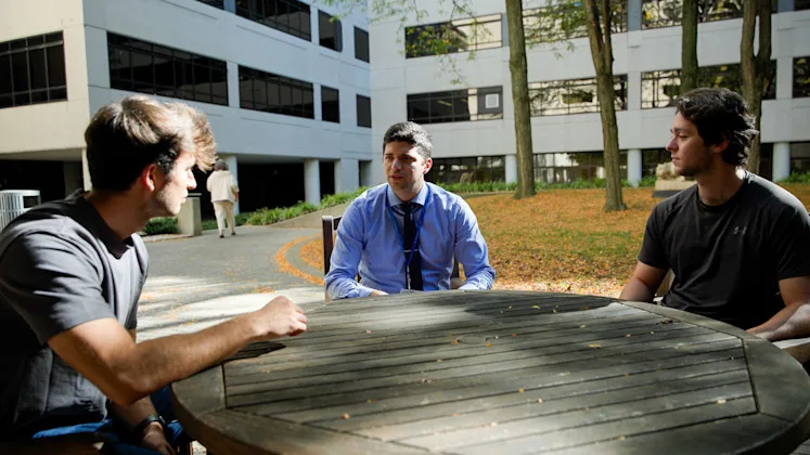 A faculty member and two students talking around an outdoor table outside the College of Medicine