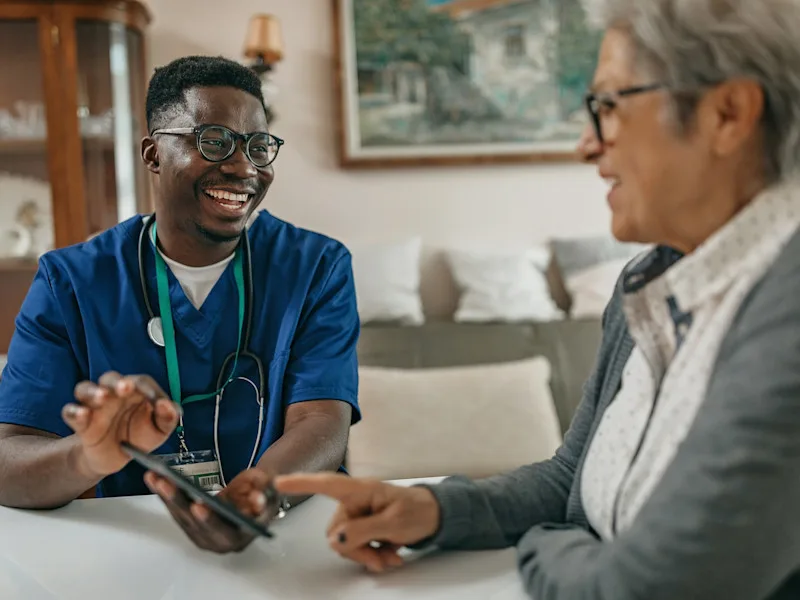 A man with a stethoscope and ID badge around his neck holds a device while talking to a woman in her home