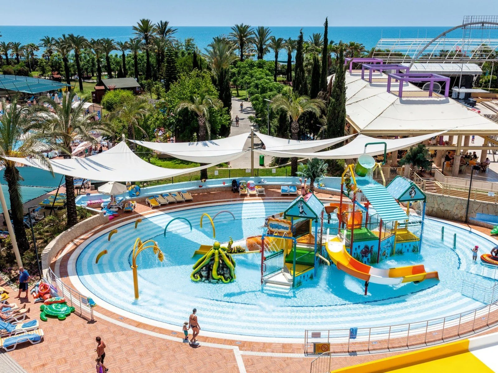 Colorful children's splash pool with slides, water features and shade sails, surrounded by palm trees near the sea