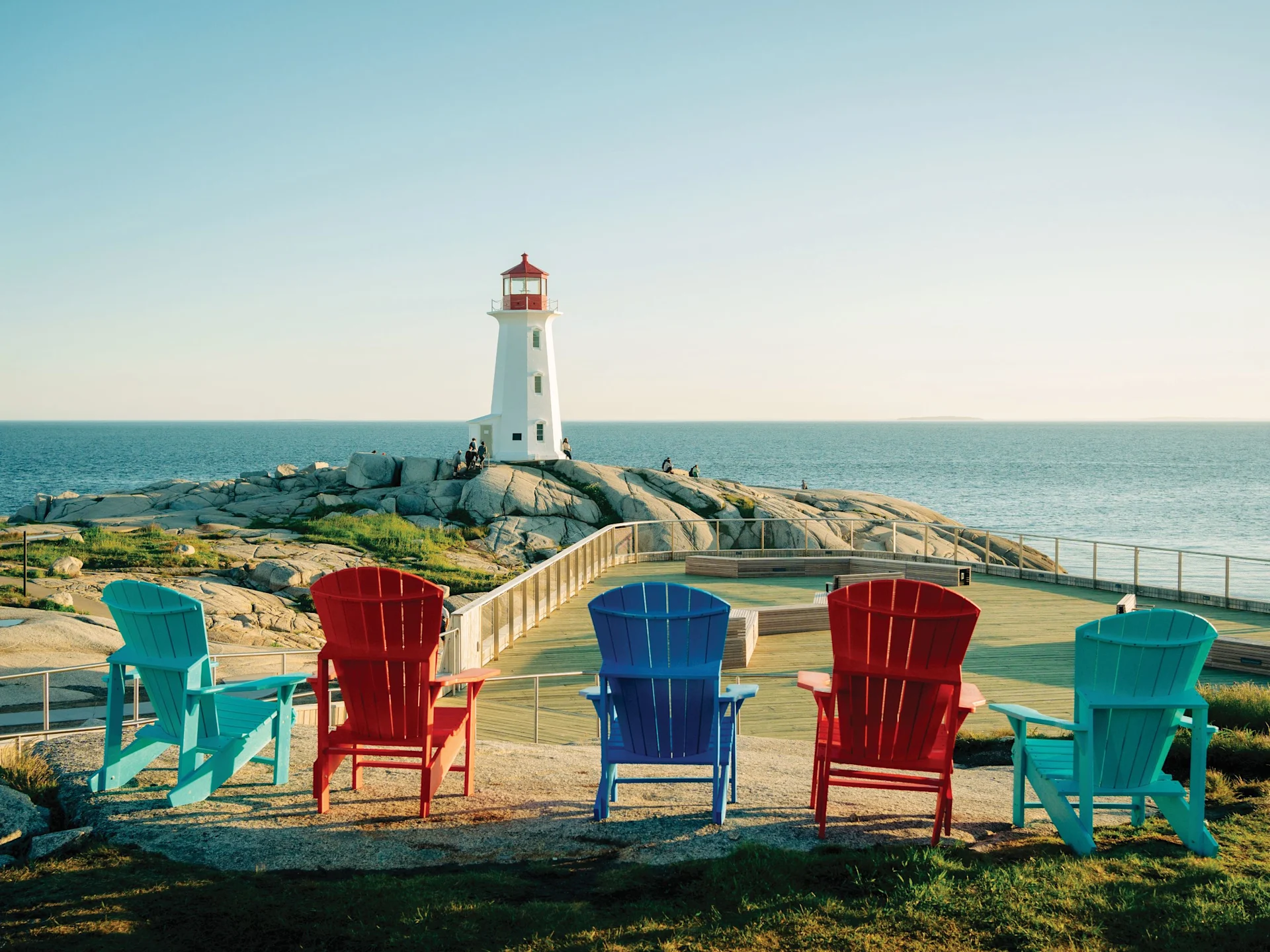 Five colorful Adirondack chairs face a white lighthouse on a rocky shore beside the sea