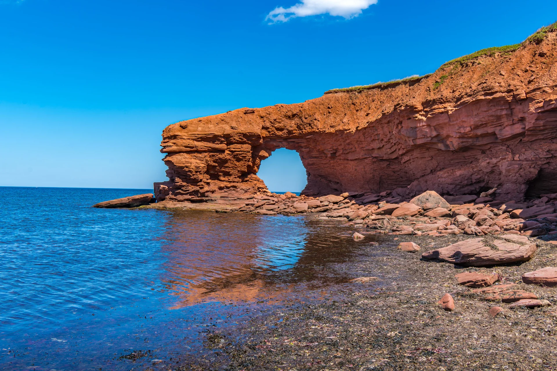 Red sandstone sea arch on a rocky shoreline beside calm blue water under a clear sky