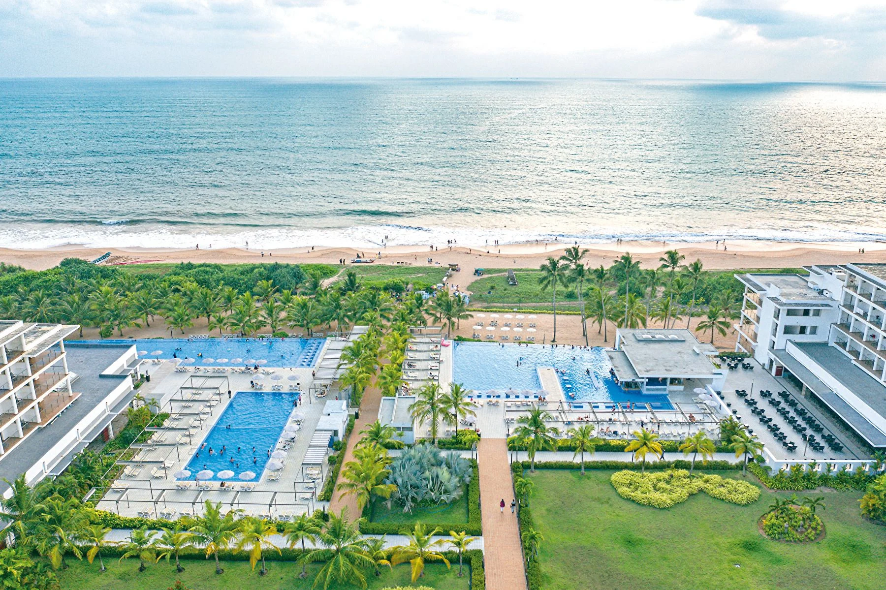 Aerial view of a beachfront resort with pools, palm trees, and ocean shoreline stretching into the distance