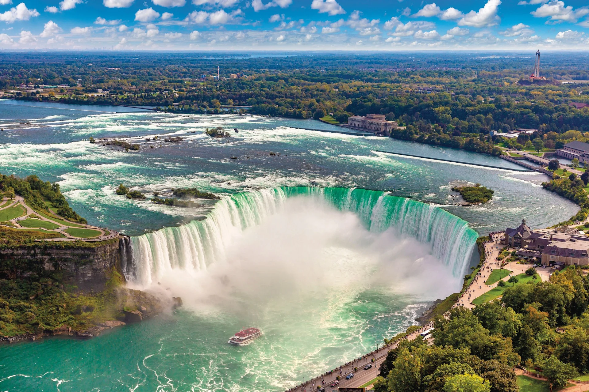 Aerial view of Niagara Falls with cascading water, rising mist, tour boat, and surrounding greenery under clouds