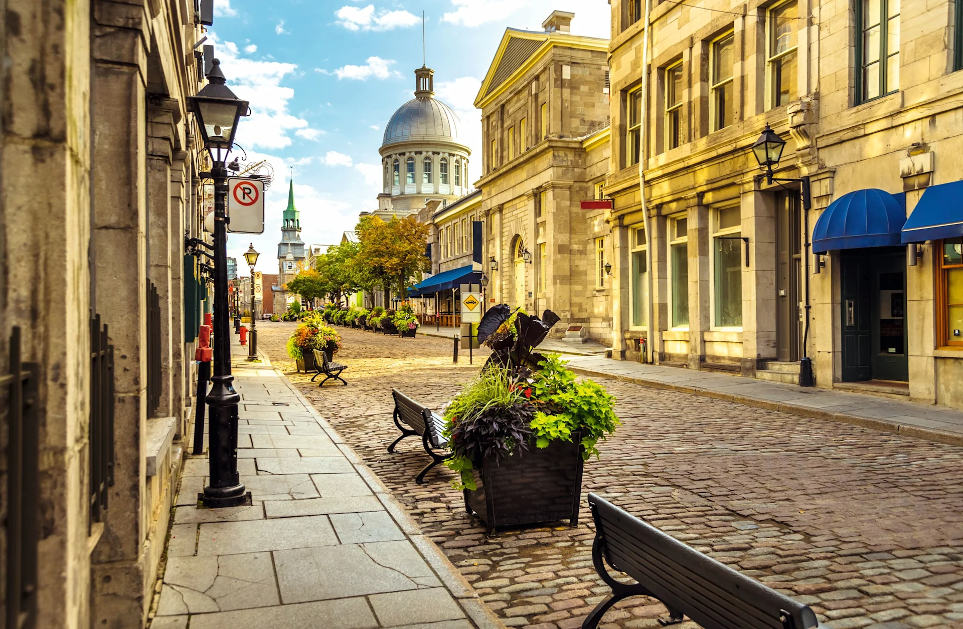 Cobblestone street with historic stone buildings, flower planters, and a domed building under blue skies.