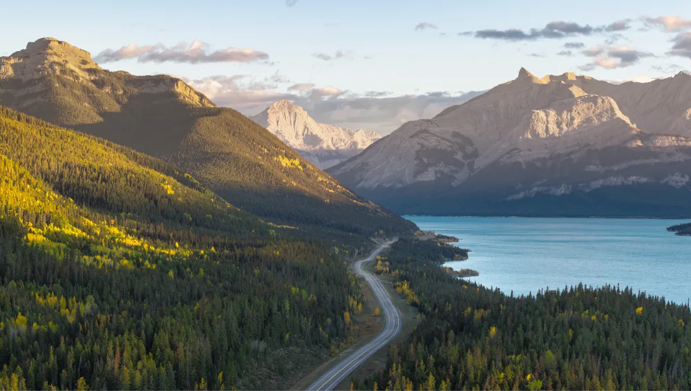 Mountain road winding through forest beside a blue lake, with sunlit peaks in the background.