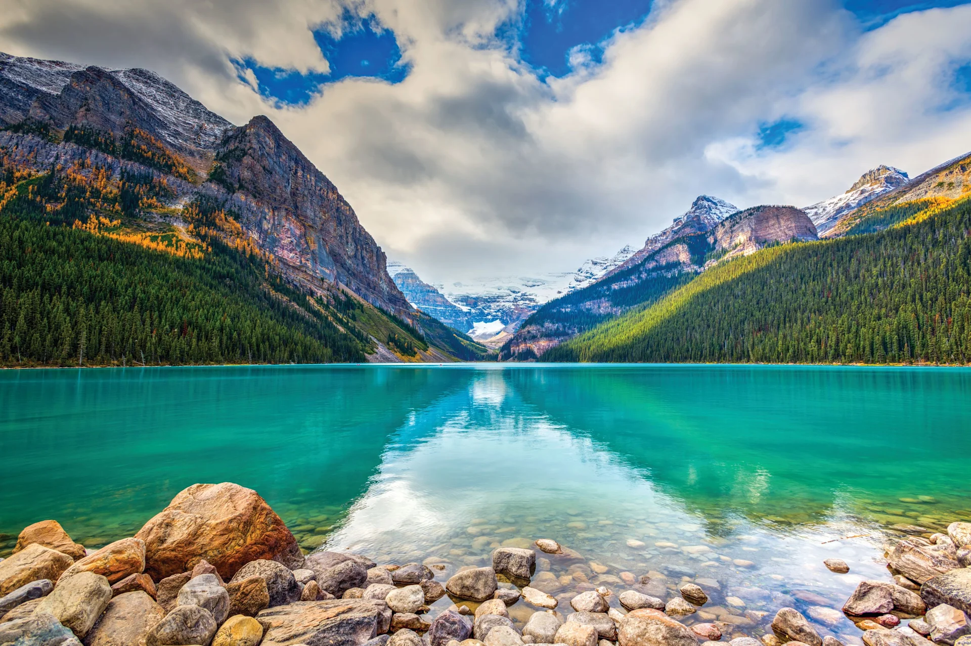 Turquoise lake with snow-capped mountains and forested shores under a partly cloudy sky.