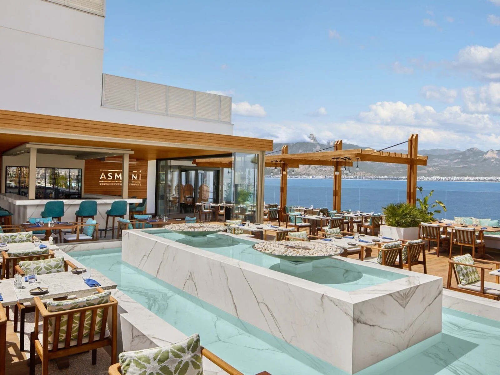 Asmani Restaurant terrace with marble water feature, wooden seating, and panoramic sea and mountain views