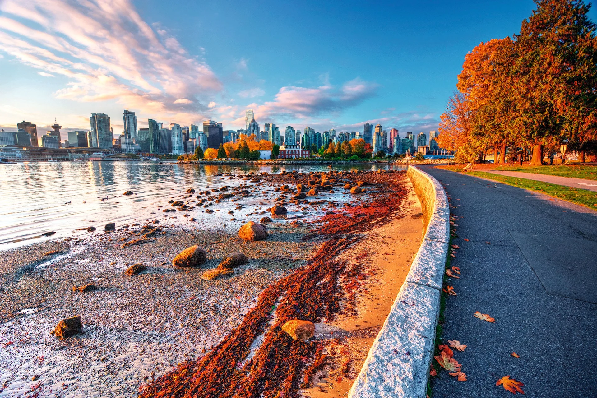 Autumn waterfront promenade with a pebble beach, water, and the Vancouver skyline in the background.