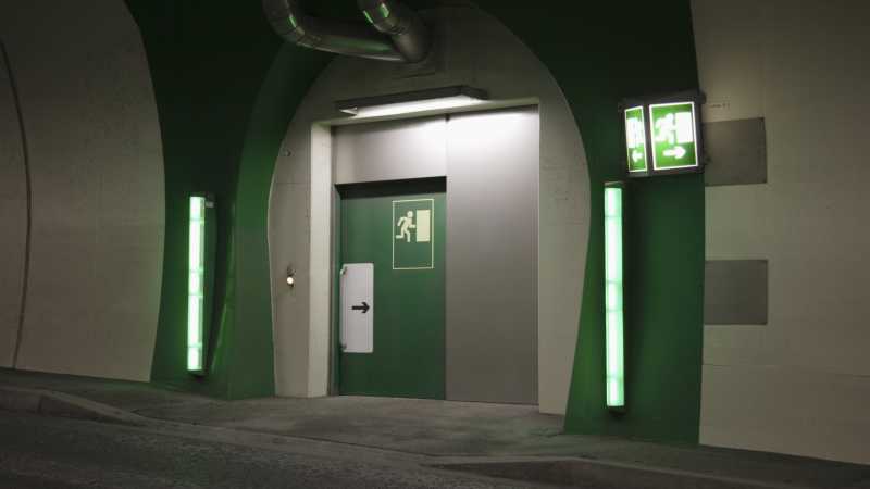 Ein grün gekennzeichneter Notausgang eines Tunnels. An den Tunnelwänden führen Notausgangsschilder mit Pfeilen zur nächsten sicheren Tür.