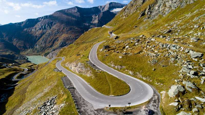 Das Bild zeigt eine Kehre einer Serpentinenstraße mit Bergen im Hintergrund.