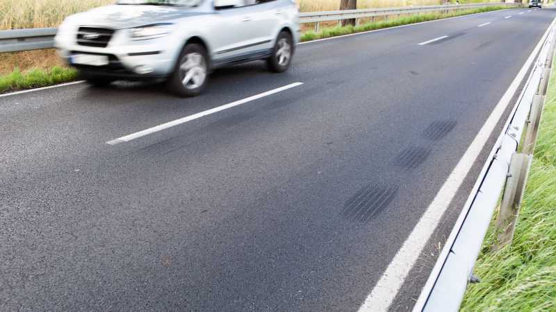 Das Bild zeigt Bremsspuren auf einer Landstraße von Nahem, während im Hintergrund ein Auto vorbeifährt.