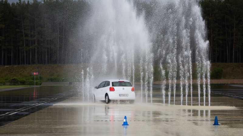 Ein weißes Auto fährt durch eine hohe Wasserfontäne. Man sieht, wie die Bremsleuchten des Wagens aufleuchten. 