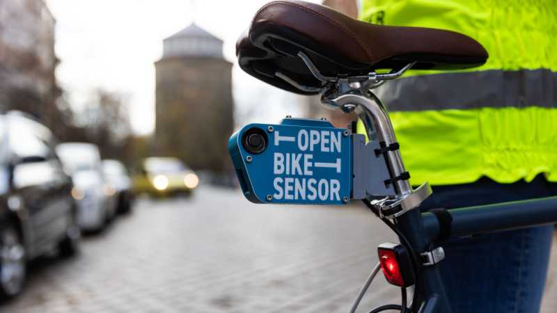 Unter dem Sattel eines Fahrrads ist ein kleiner blauer Kasten montiert. Es ist der „OpenBikeSensor“. Im Hintergrund ist der Wasserturm in Prenzlauer Berg in Berlin zu sehen.