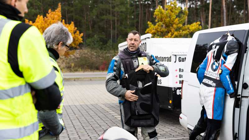 Trainer Peer spricht mit den beiden Teilnehmenden und hält Motorradkleidung in der Hand.