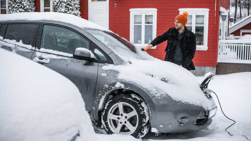 Ein Mann entfernt Schnee von seinem Elektroauto, das an einen Ladestecker angeschlossen ist.