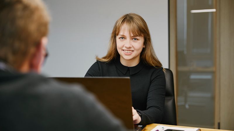 Portrait von Madlin Schulz. Sie sitzt an einem Tisch vor einem Laptop und blickt lächelnd in die Kamera. 