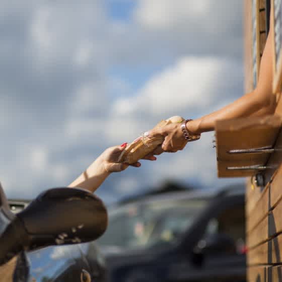 Man sieht eine Hand, die aus einem Drive-Through-Fenster einer anderen Person in einem Auto Essen reicht.