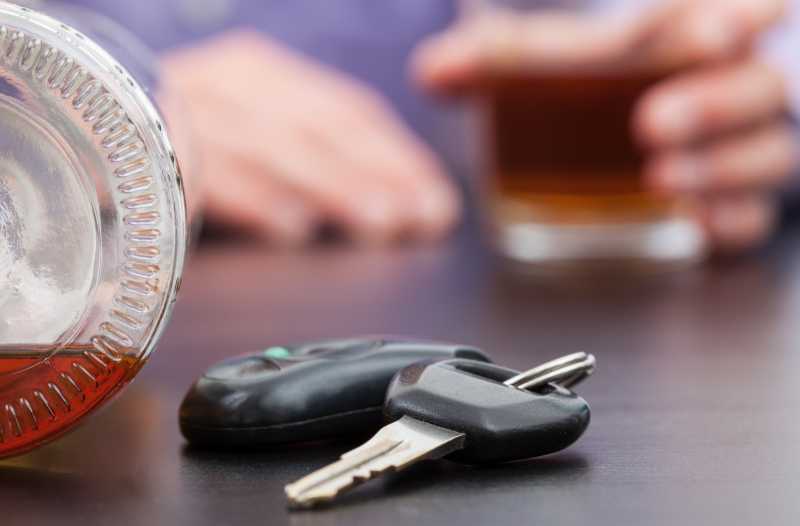 Man sieht ein Paar Autoschlüssel und eine liegende Flasche mit brauner Flüssigkeit, während im Hintergrund jemand ein volles Alkoholglas in seiner Hand hält.