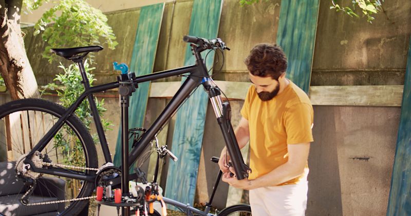Ein Mann mit Bart in einem gelben T-Shirt repariert im Freien ein schwarzes Fahrrad, das auf einem Montageständer befestigt ist. Er arbeitet konzentriert an der Fahrradgabel.
