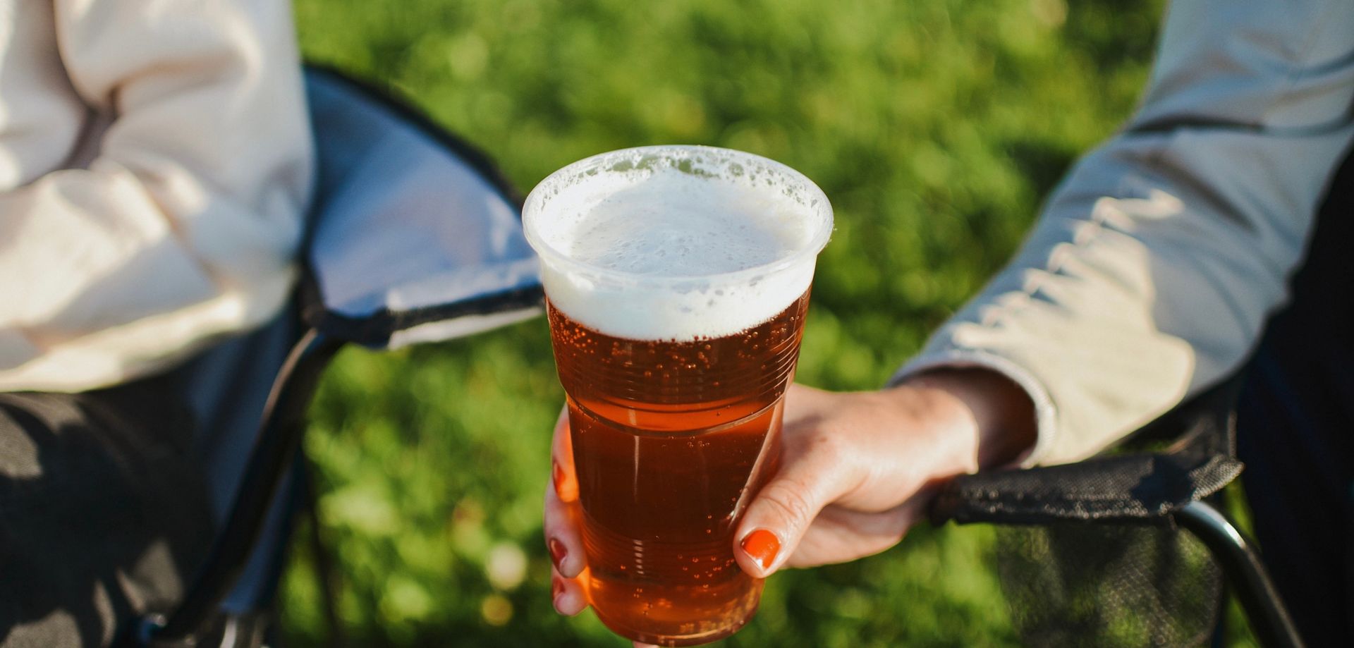 Nahaufnahme einer Hand, die einen Becher bernsteinfarbenes Bier hochhält. Im unscharfen Hintergrund sitzen zwei Personen im Sonnenlicht auf Klappstühlen im Gras.
