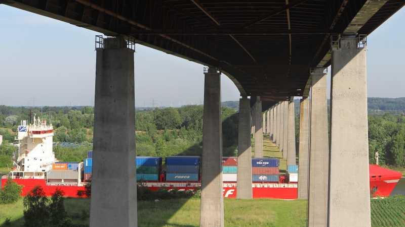 Das Bild zeigt ein Containerschiff, das unter einer hohen Brücke durchfährt.