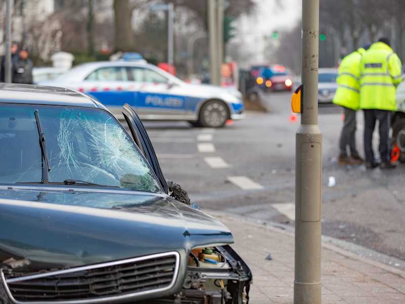 Vorne im Bild ist ein beschädigtes dunkles Auto mit zerbrochener Windschutzscheibe und offener Motorhaube abgebildet. Im Hintergrund sind ein Polizeiauto.