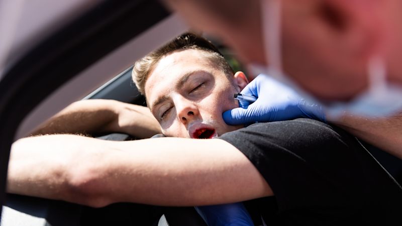 Ein Verletzter liegt über dem Lenkrad. Aus seinem Mund tropft eine rote Flüssigkeit. Ein Sanitäter untersucht ihn. 