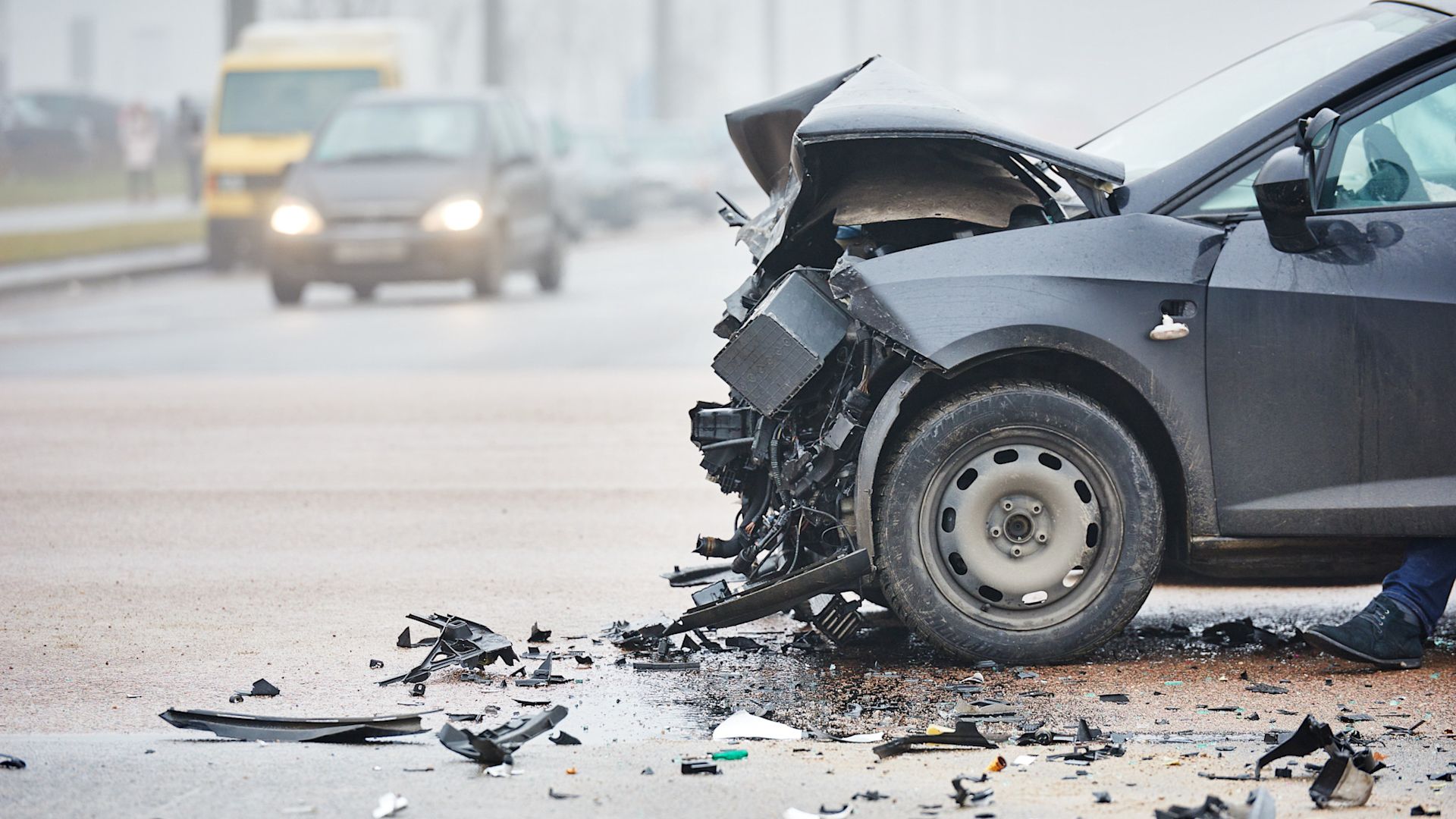 Ein schwarzes Auto mit stark beschädigter Front steht nach einem Unfall auf einer nassen Straße. Überall liegen Trümmerteile verstreut, und im Hintergrund sind verschwommen weitere Fahrzeuge zu sehen.