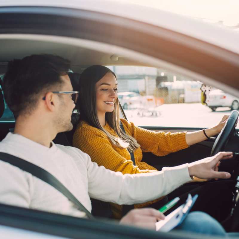 Ein Fahrlehrer und eine Frau sitzen im Auto. Der Fahrlehrer zeigt der Frau am Steuer ein technisches Detail.