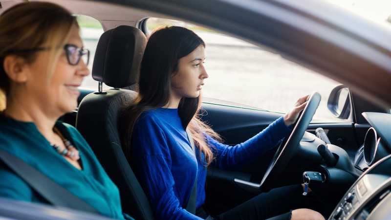 Ein junges Mädchen mit dunklen Haaren und blauem Pullover sitzt am Lenkrad eines Autos. Neben ihr sitzt eine blonde Frau mit Brille und lächelt. 