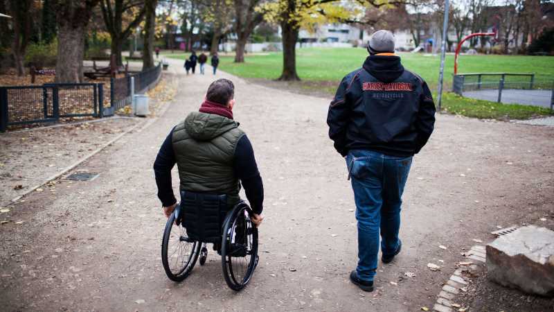 zwei Männer gehen im Park spazieren, einer sitzt im Rollstuhl und hat keine Beine.