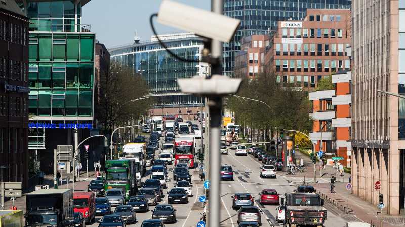 Man sieht eine vielbefahrene Straße in der Innenstadt von Hamburg, an dessen Seiten hohe Gebäude stehen und die von Verkehrsbeobachtungskameras überwacht wird.