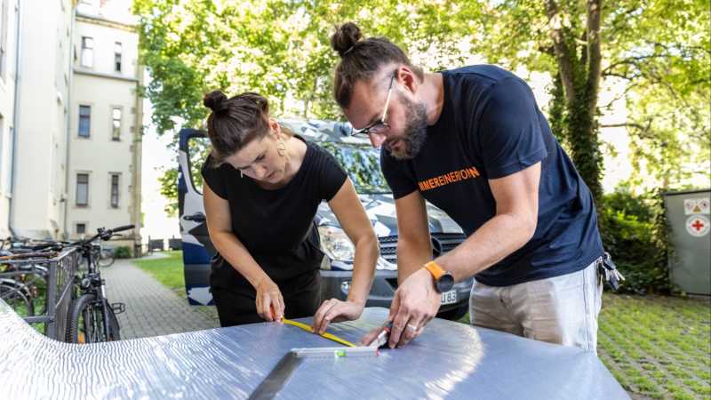 Eine Frau und ein Mann stehen an einer Arbeitsplatte und schneiden Isoliermaterial. Im Hintergrund sieht man einen Bus, der im Hinterhof steht.