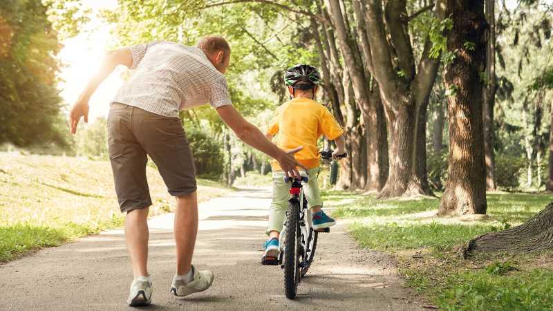 ein Mann hilft einem Kind auf einem Fahrrad beim Anfahren.