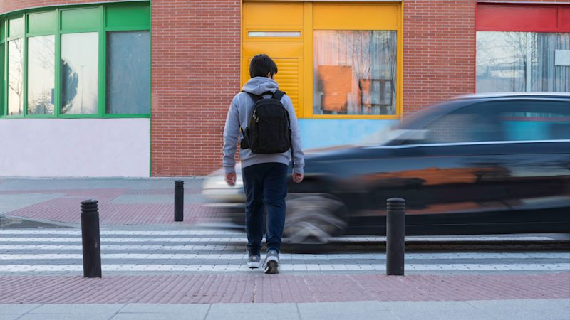 Ein Jugendlicher mit Rucksack überquert einen Zebrastreifen in der Stadt, während ein schwarzes Auto den Fußgängerüberweg missachtet und schnell an ihm vorbeifährt.
