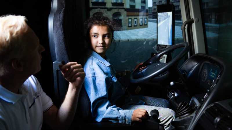 Ein Mädchen sitzt am Lenkrad eines Lkw-Simulator. Dabei bekommt es eine Erklärung eines Trainers.  