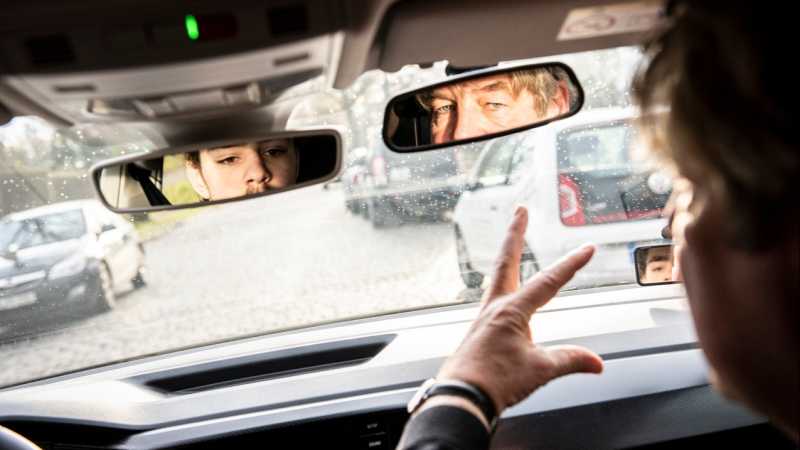 Man sieht die Gesichter von Fahrlehrer und Fahrschüler in den zwei Rückspiegeln des Fahrschulautos.