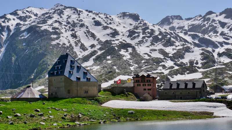 Das Bild zeigt Restaurants an einem Bergsee am Gotthardpass mit schneebedeckten Bergen im Hintergrund.