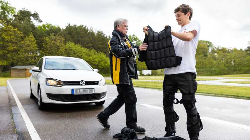 Ein junger Mann hält eine Weste. Ein älterer Mann hilft ihm diese anzuziehen. Im Hintergrund steht ein weißes Auto. 