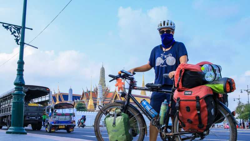 Luisa Rische steht mit ihrem Fahrrad in Thailand vor einer vielbefahrenen Straße mit einem Tempel im Hintergrund.