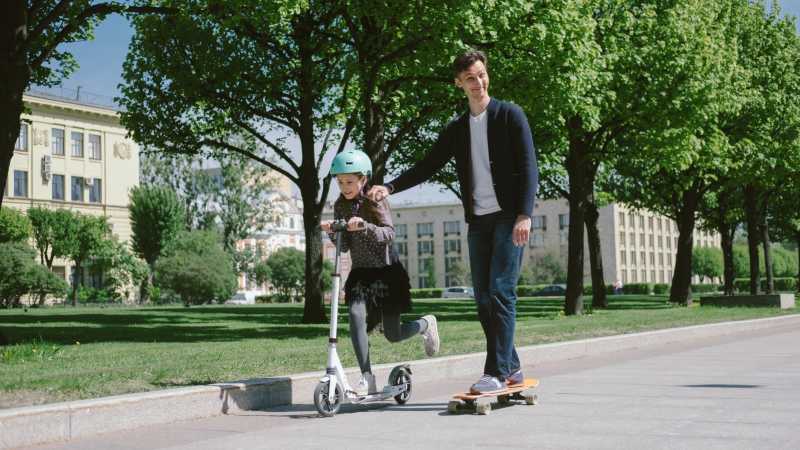 Ein Mann fährt auf einem Skateboard einen breiten Weg im Park entlang. Er lächelt in die Kamera. Ein Mädchen fährt neben ihm auf einem Tretroller. Sie trägt einen Sicherheitshelm.