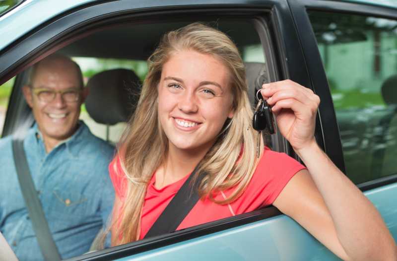 Das Bild zeigt eine junge Fahrerin am Steuer eines Autos. Sie sitzt neben ihrem Vater und hält den Autoschlüssel glücklich in die Kamera.