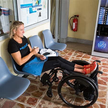 Kirsten Bruhn sitzt auf einer Reihe Stühle und hat ihre Füße auf ihren Rollstuhl gelegt.