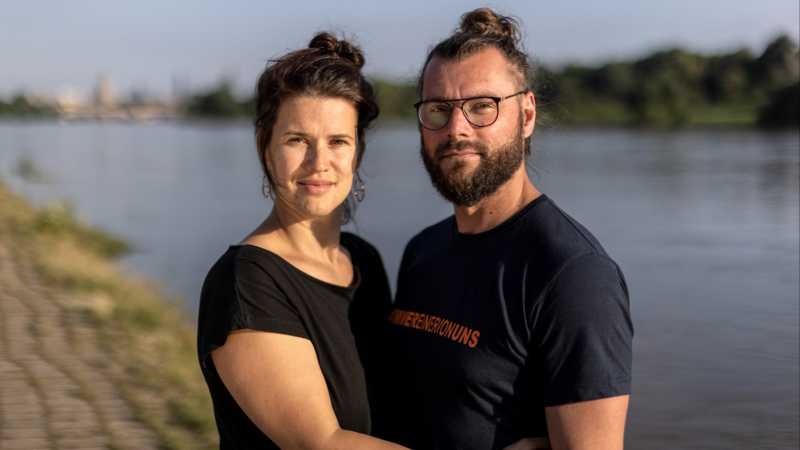 Eine Frau und ein Mann lachen in die Kamera. Zu sehen sind Katja Wiedemann und Torsten Grimm. Im Hintergrund sieht man die Elbe. 