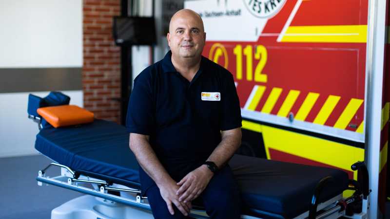 Ein Mann sitzt auf einer Krankentrage und lächelt in die Kamera. Im Hintergrund sieht man einen Rettungswagen. Es ist Sven Baumgarten. 