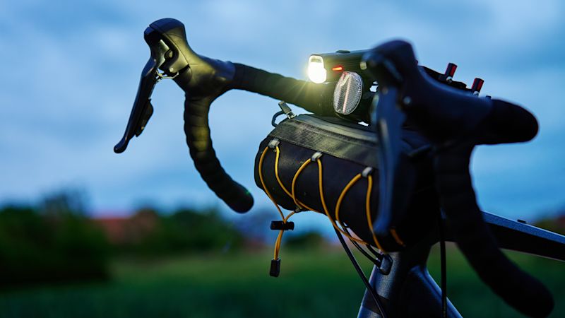 Nahaufnahme eines Fahrradlenkers bei Dämmerung. Der Lenker ist mit einer leuchtenden Frontlampe und einem Reflektor ausgestattet. Darunter befindet sich eine schwarze Fahrradtasche mit gelben Kordeln, die am Rahmen befestigt ist. Im Hintergrund sind unscharfe grüne Wiesen und ein bewölkter Himmel zu sehen.