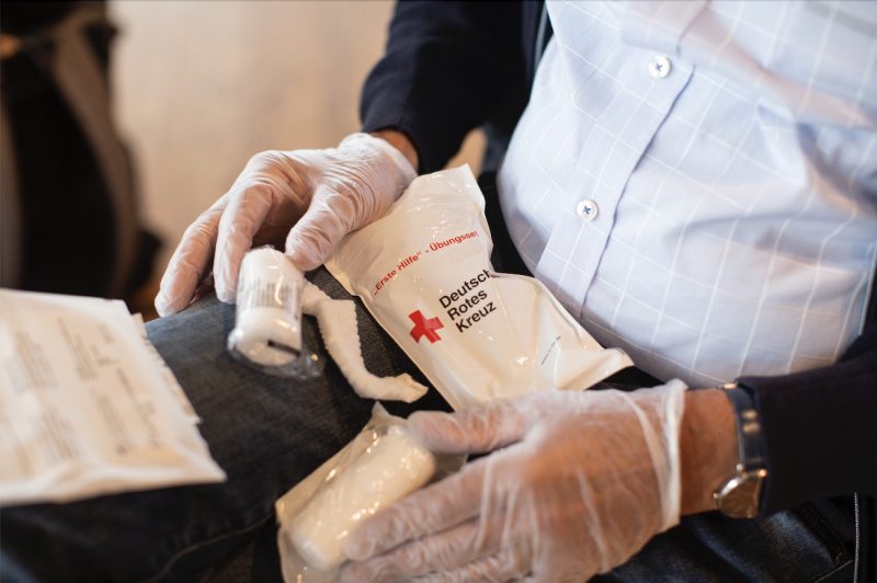 Ein Mann trägt Plastikhandschuhe und hat verpackte Verbände des Deutschen Roten Kreuzes im Schoß.