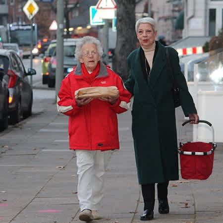 zwei ältere Frauen laufen eine Straße entlang, die jüngere trägt einen Einkaufskorb.