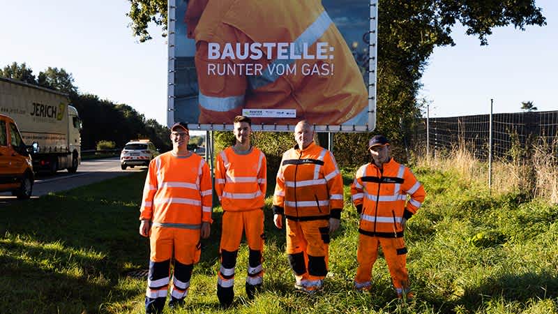 Vier Straßenwärter in orangefarbener Schutzkleidung stehen vor einem Autobahnplakat.